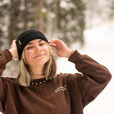 Woman standing in a snowy forest wearing a black beanie with a yellow tab with Nalle on it.