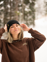 Woman standing in a snowy forest wearing a black beanie with a yellow tab with Nalle on it.