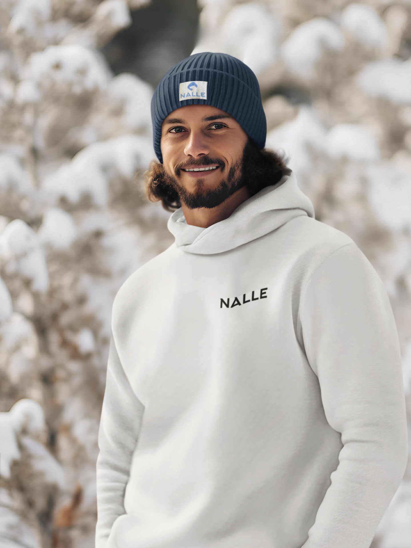 Man wearing a blue Nalle beanie and gray Nalle hoodie standing in front of a snow laden tree.