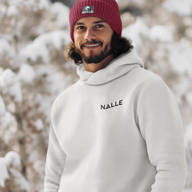 Man wearing a red Nalle beanie and gray Nalle hoodie standing in front of a snow laden tree.