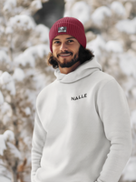 Man wearing a red Nalle beanie and gray Nalle hoodie standing in front of a snow laden tree.