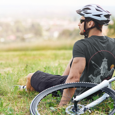 Man sitting on the grass in front of his bike wearing a bike helmet and black Nalle tee with the Mooka wizard on it.