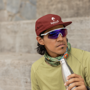 Man wearing a red cap with the Nalle logo on it sitting in front of a wall resting after a hike drinking from a metal water bottle.